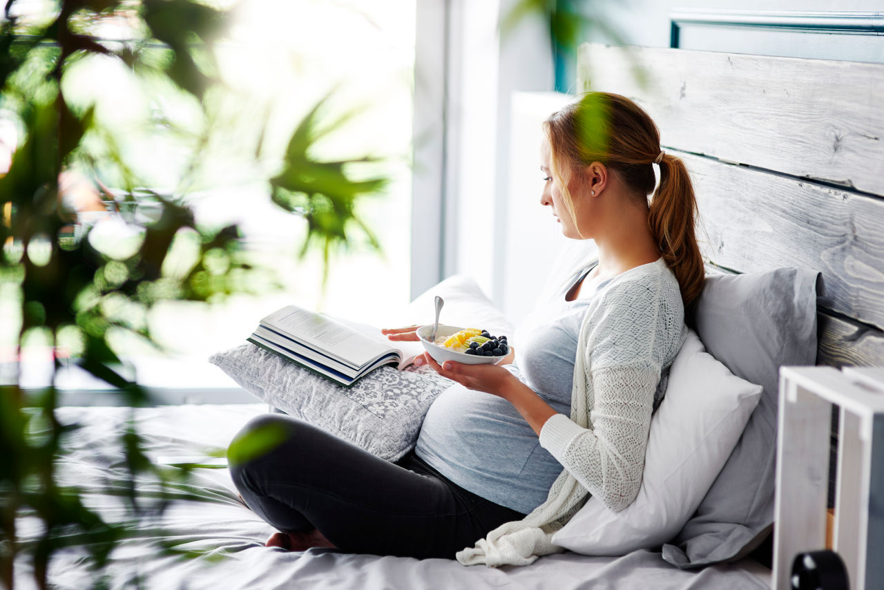 Frau schwanger sitzt auf Sofa mit Buch und isst Früchte aus Schüssel